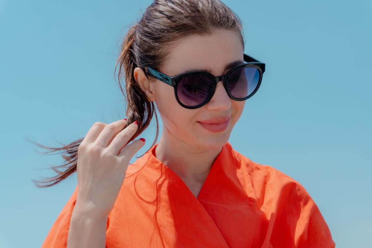 A woman wearing sunglasses and an orange blouse enjoys a sunny day outdoors.