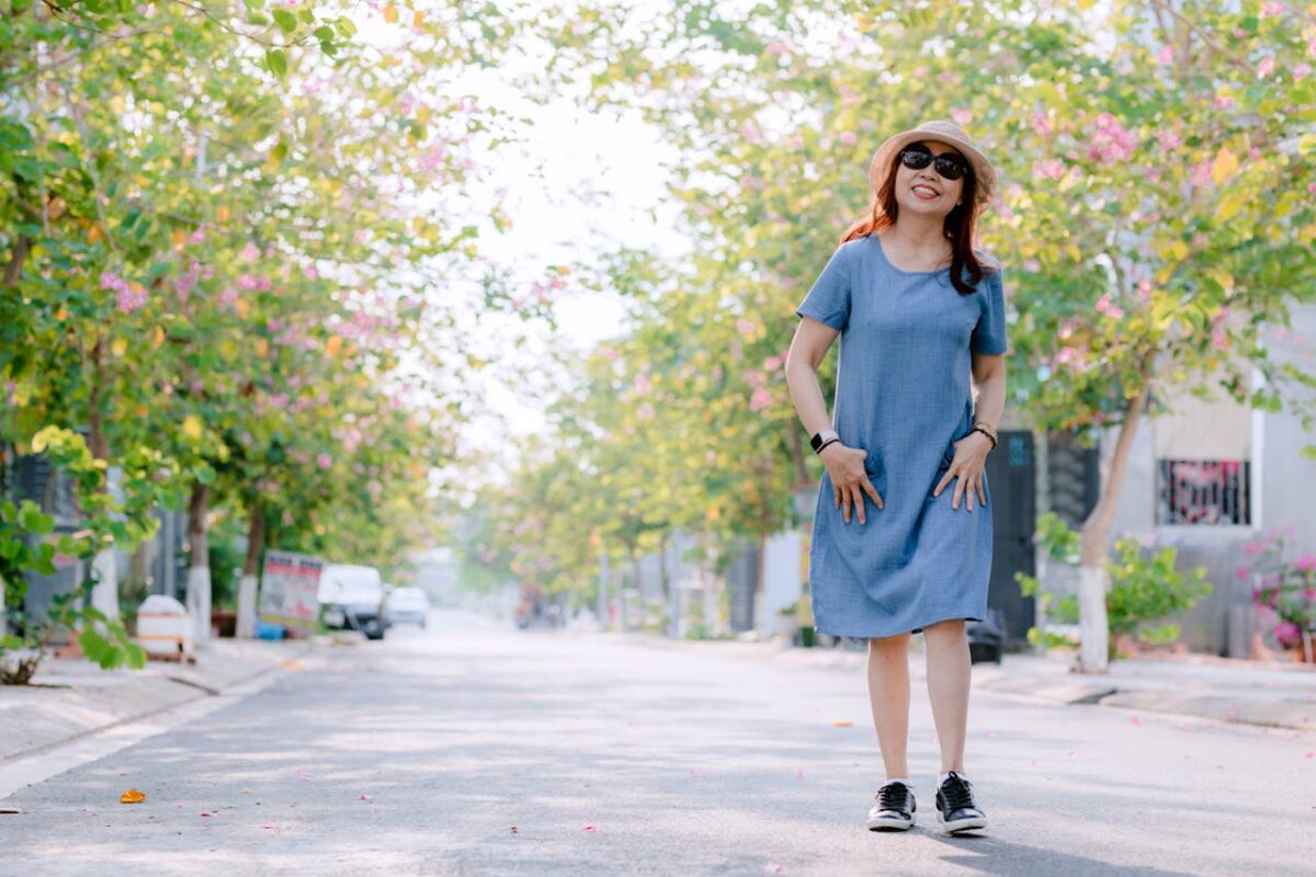 Smiling woman in a blue dress and hat standing on a sunny, tree-lined street.